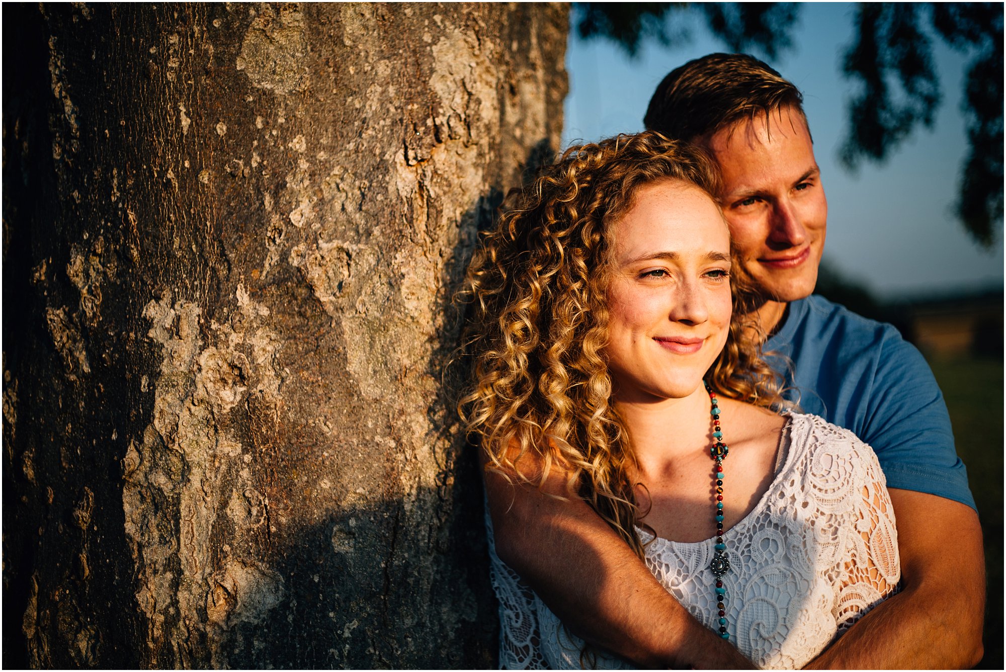 Warm lighting on engaged couple as they lean against large tree watching sunset