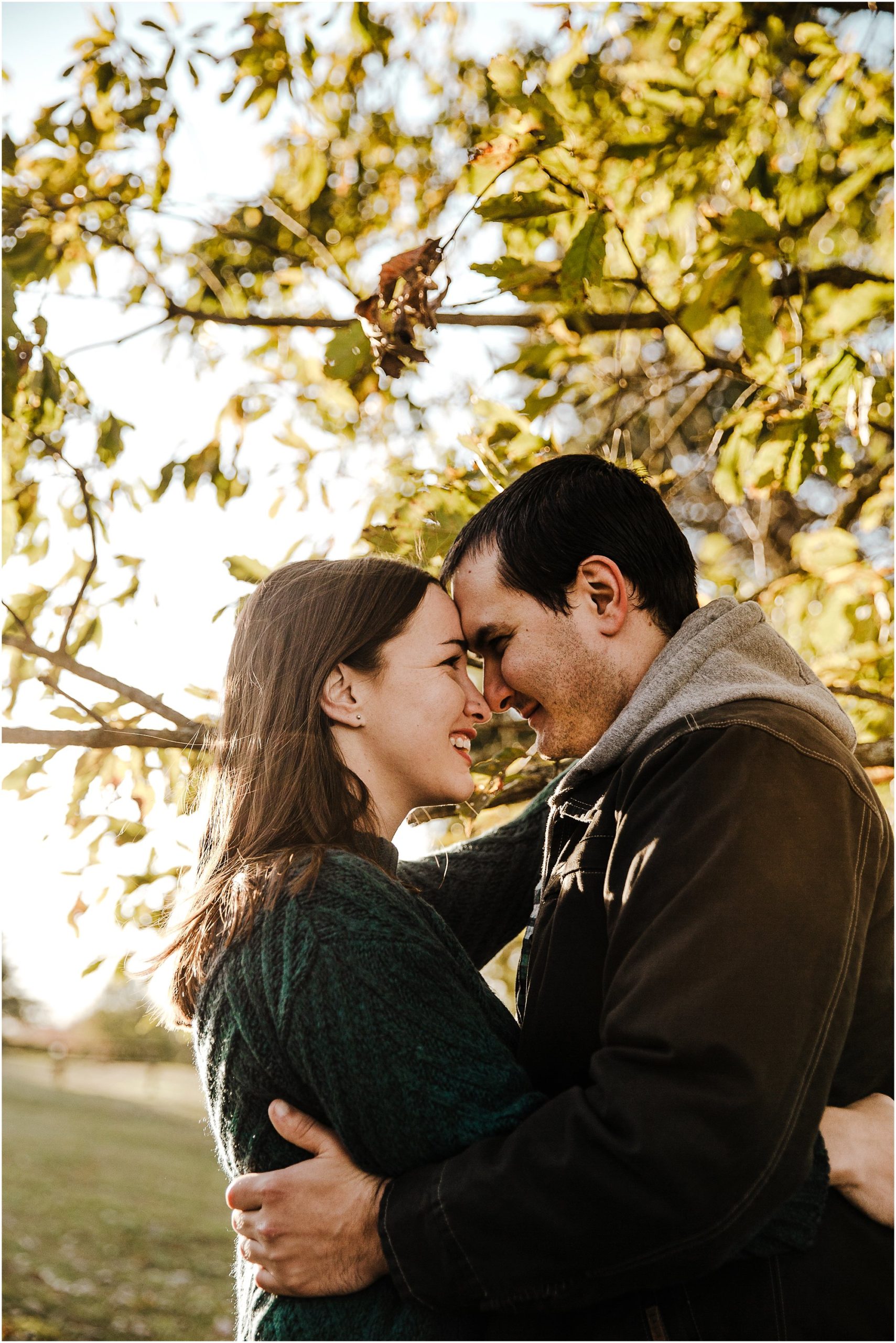 Engaged couple with their foreheads together giggling in front of backlit fall leaves.